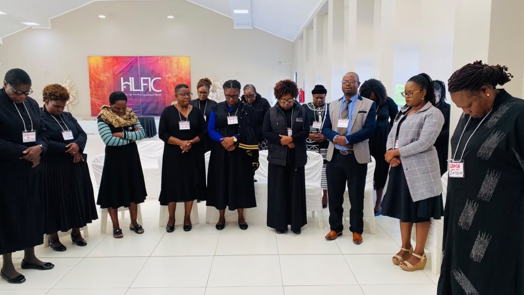 Participants stand in a circle with heads bowed in prayer in a hall.