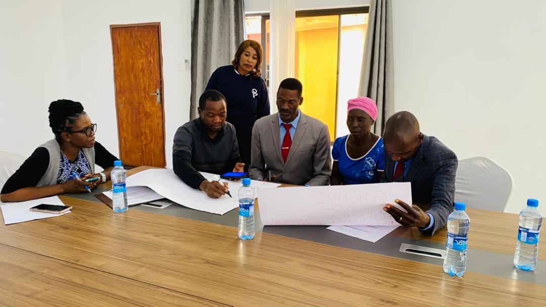 A group gathered around a boardroom table, one participant writing on a large sheet while the others watch.